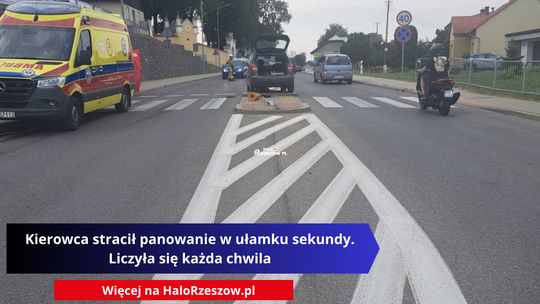 Na zdjęciu widać miejsce wypadku drogowego. Na pierwszym planie, po lewej stronie, stoi żółta karetka pogotowia. W głębi, na środku jezdni, stoi samochód osobowy z otwartym bagażnikiem, a obok niego, na poboczu, widać uszkodzone elementy