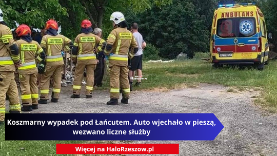 Miejsce wypadku drogowego w Husowie pod Łańcutem. Po lewej stronie stoi grupa strażaków w mundurach bojowych Państwowej Straży Pożarnej i OSP. Po prawej stronie na trawiastym poboczu zaparkowana jest żółta karetka pogotowia ratunkowego z włączonymi sygnałami świetlnymi i napisem Ambulans.