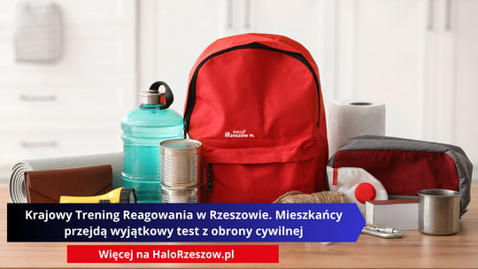 Czerwony plecak otoczony przedmiotami pierwszej potrzeby: butelką wody, puszkami jedzenia, latarką, apteczką, maską ochronną i ręcznikiem papierowym, leżącymi na drewnianym blacie.