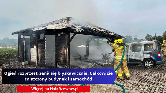 Na zdjęciu widać strażaków gaszących pożar drewnianego, otwartego garażu, pod którym znajduje się mocno uszkodzony samochód dostawczy. Z pogorzeliska unosi się gęsty dym