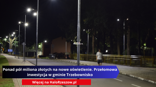 Nocne zdjęcie przedstawiające oświetloną ulicę i chodnik z rzędem wysokich, nowoczesnych latarni ulicznych. W tle widać drzewa oraz postać pieszego spacerującego poboczem.