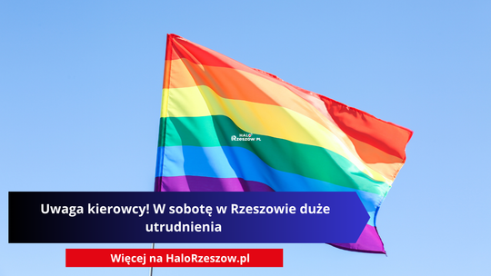 Tęczowa flaga symbolizująca społeczność LGBT na tle czystego, błękitnego nieba z nałożonym napisem ostrzegawczym dla kierowców o utrudnieniach w Rzeszowie.