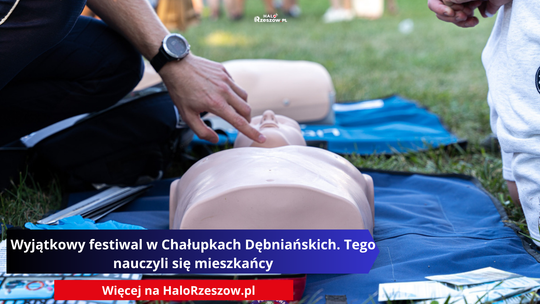 Zbliżenie na męską dłoń instruktora wskazującą punkt do ucisku na klatce piersiowej fantomu medycznego leżącego na niebieskiej macie na trawie. W tle widać inne osoby. Na grafice nałożono fioletowy baner z napisem: "Wyjątkowy festiwal w Chałupkach Dębniańskich. Tego nauczyli się mieszkańcy".