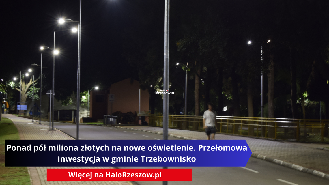 Ponad pół miliona złotych na nowe oświetlenie. Przełomowa inwestycja w gminie Trzebownisko Nocne zdjęcie przedstawiające oświetloną ulicę i chodnik z rzędem wysokich, nowoczesnych latarni ulicznych. W tle widać drzewa oraz postać pieszego spacerującego poboczem.